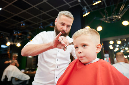 Man barber cutting little boys hair using comb and scissors. Child getting haircut from adult male, likely barber. Professional hairdresser and cute client at modern barbershop.の写真素材