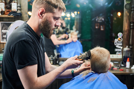 Professional barber concentrated on his work. Little kid getting first haircut in barbershop. Male hairdresser using electric shaver to cut boys hair.の写真素材