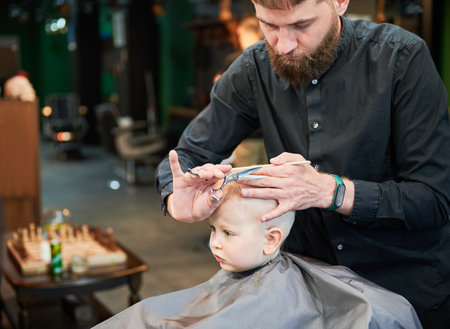 Man barber cutting little boys hair using comb and scissors. Child getting haircut from adult male, likely barber. Professional hairdresser and cute client at modern barbershop.の写真素材