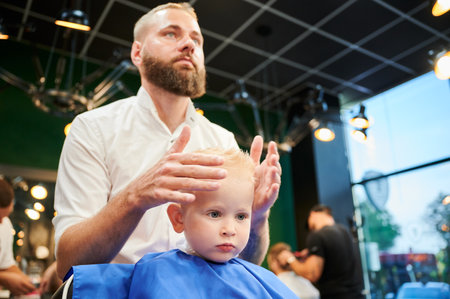 Young child getting haircut in modern barbershop. Hairdresser making final touches in new haircut, shaping childs hair by hands.の写真素材