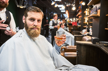 Handsome man enjoying his drink in barbershop. Male customer relaxing while getting a haircut. Confident man holding glass and looking at camera.の写真素材