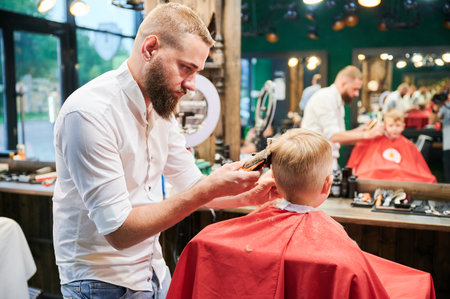 Barber using comb and shaver to cut hair. Professional hairdresser shaving little kids nape.の写真素材