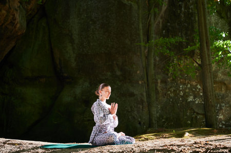 Woman practicing yoga outdoors in forest. Barefoot female on yoga mat surrounded by trees and large rocks, which suggests peaceful, natural environment ideal for meditation or yoga practice.の写真素材