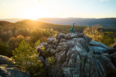 Woman practicing yoga and meditation on the top of huge boulder in the evening. Beautiful sunset, autumn forests, rocks and hills on background. Aerial drone viewの写真素材