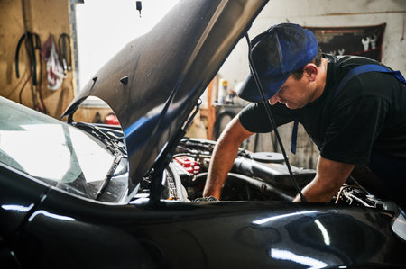 Worker, repairman wearing cap, repairing car engine. Hardworking man opening car hood, looking, working in garage, auto, servicing center. Concept of manual labor.の写真素材