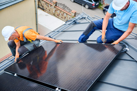 Workers building photovoltaic solar panel system on rooftop of house. Men technicians in helmets and gloves installing solar module with help of hex key outdoors. Alternative energy generation conceptの写真素材