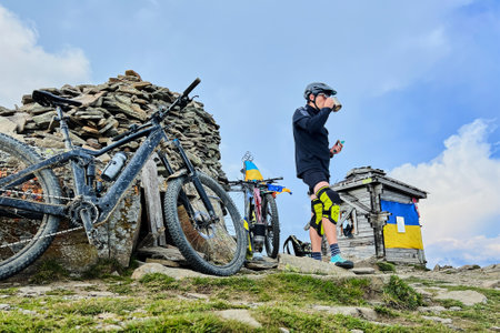 Cyclist man resting on the top of hill while riding electric bike outdoors. Male tourist drinking tea, enjoying beautiful mountain landscape, wearing helmet. Concept of active leisure.の写真素材