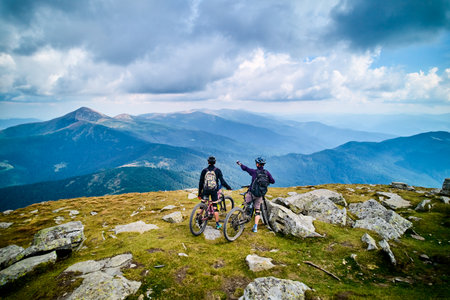 Two cyclists men riding electric bikes outdoors. Back view of male tourists resting on the top of hill, enjoying beautiful mountain landscape, wearing helmet and backpack. Concept of active leisure.の写真素材