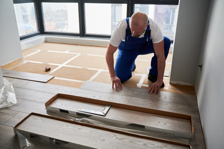 Man in work overalls installing laminate timber flooring in room with window. Male construction...
