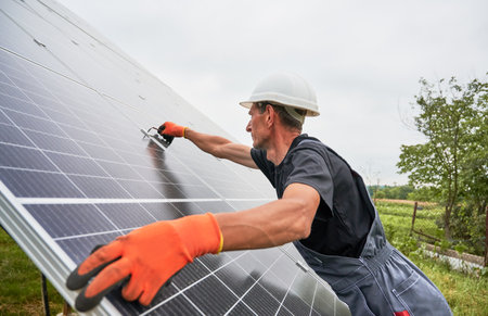 Male worker in safety helmet checking middle clamp while mounting solar photovoltaic panel system. Man in work gloves installing solar modules in grassy field with white sky on background.の写真素材