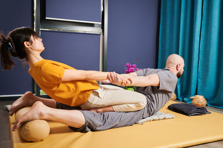 Masseuse making thai yoga massage. Woman therapist doing traditional massage treatment, stretching male patients body. Bald client lying on mat, enjoying hand massage in calm atmosphere.の写真素材