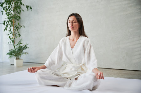 Beautiful woman in white dress sitting on mat in lotus position before Thai massage, practicing yoga.の写真素材