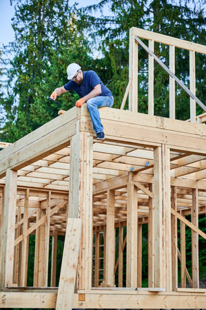 Carpenter building wooden, two-story frame house near woods. Bearded man with spectacles hammering nail while wearing protective helmet. The concept of contemporary ecological construction.の写真素材