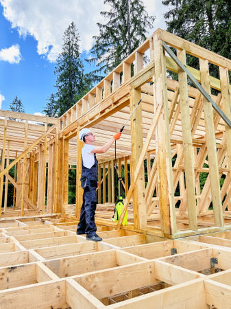 Laborer constructing wooden frame house near forest. Man treating woods, applying fire retardant using sprayer, while dressed in protective suit, helmet. Concept of modern eco-friendly construction.の写真素材