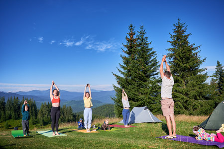Group of people doing yoga pose outdoor in camping in the mountains. Adults and children standing on yoga mats, each doing yoga pose under clear blue sky in morning.の写真素材