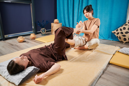 Masseuse making thai yoga massage. Woman therapist doing traditional massage treatment for female patient indoors. Client lying on mat, meditating, enjoying hand massage in calm atmosphere.の写真素材