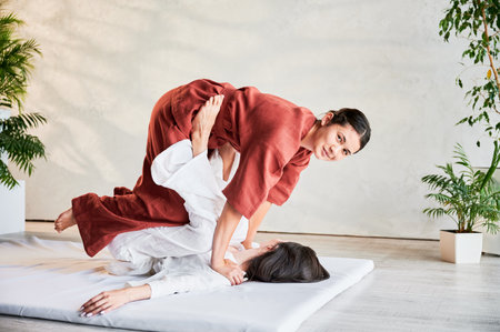Masseuse making thai yoga massage. Woman therapist doing traditional massage treatment, stretching female patients body. Client lying on mat, enjoying hand massage in calm atmosphere.の写真素材