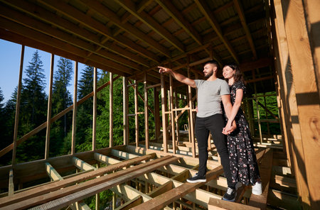 Man and woman inspecting their future wooden frame dwelling nestled in the mountains near forest. Youthful couple at construction site in early morning. Concept of contemporary ecological constructionの写真素材