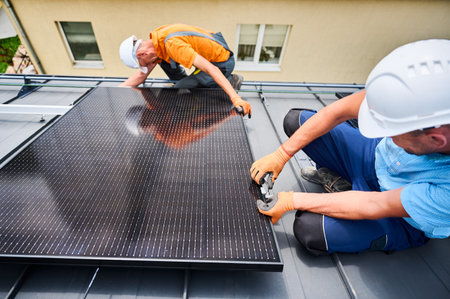 Workers building photovoltaic solar panel system on rooftop of house. Men technicians in helmets and gloves installing solar module with help of hex key outdoors. Renewable energy generation concept.の写真素材