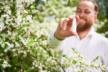 Focus on man hands holding nasal spray. Smiling man allergic using medical nasal drops, suffering from seasonal allergy in blossoming garden. Spring allergy concept.の写真素材