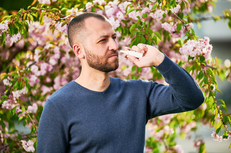Man allergic using medical nasal drops, suffering from seasonal allergy at spring in blossoming garden. Handsome man treating runny nose in front of blooming tree outdoors. Spring allergy concept.の写真素材