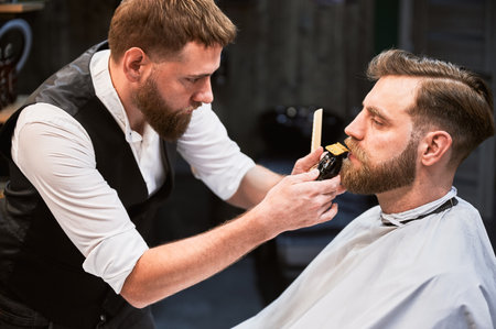 Professional hairdresser shaping thick beard. Close-up of male barber in fancy outfit shaving beard. Appointment in barbershop.の写真素材