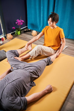 Masseuse making thai yoga massage. Woman therapist doing traditional massage treatment, stretching male patients body. Client lying on mat, enjoying hand massage in calm atmosphere.の写真素材