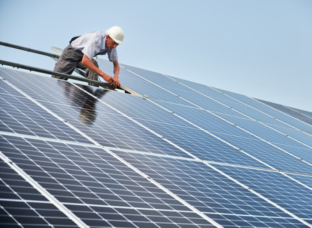 Male solar technician installing solar modules on metal construction. Man in work overalls and safety helmet mounting photovoltaic solar panels under blue sky. Concept of alternative renewable energy.の写真素材