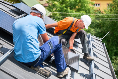 Workers building solar panel system on rooftop of house. Two men installers in helmets installing photovoltaic solar module outdoors. Alternative, green and renewable energy generation concept.の写真素材