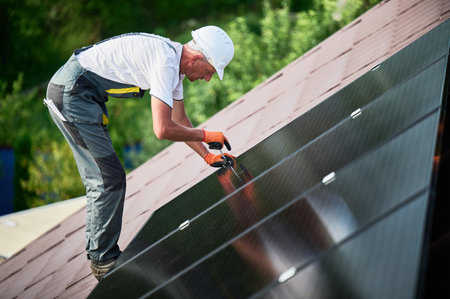 Worker building photovoltaic solar panel system on rooftop of house. Man engineer in helmets and gloves installing solar module with help of hex key outdoors. Renewable energy generation concept.の写真素材