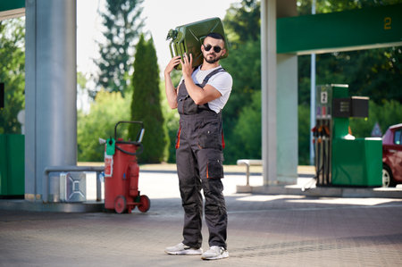 Strong worker holding canister full of fuel on shoulder. Confident gas station operator caring heavy cisterns with gas. Male worker in sunglasses and overalls standing on background of gas station.の写真素材