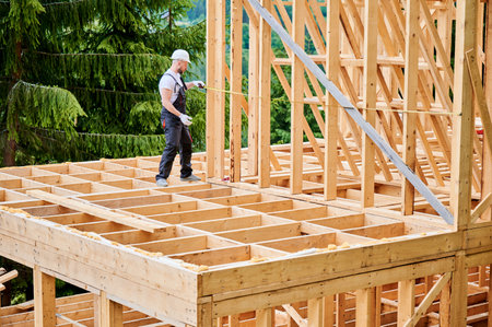 Carpenter constructing house with wooden frame. Man measures distance using tape measure, wearing protective clothing and helmet. Concept of environmentally sound, modern construction.の写真素材