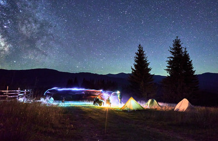 Blurred silhouettes of hikers resting around campfire, surrounded by illuminated tents under...