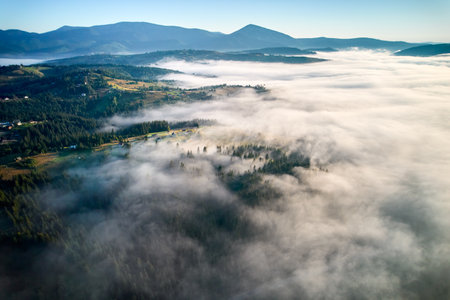 Aerial view reveals misty morning landscape with rolling hills and villages. Golden sunlight breaks through fog, illuminating patches of green and creating dreamy, ethereal scene under clear blue sky.の写真素材