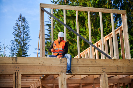 Carpenter constructing two-story wooden frame house near forest. Bearded man hammering nails into...
