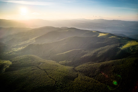 Aerial view of serene mountain landscape at sunset. Forested hills bathed in warm, golden light, creating tranquil atmosphere. Rolling hills stretch into the distance, covered in dense, lush greenery.の写真素材