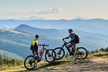 Two cyclists men riding electric bikes outdoors. Smiling male tourists resting on the top of hill, enjoying beautiful mountain landscape, wearing helmet and backpack. Concept of active leisure.の写真素材
