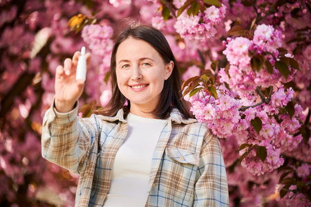 Woman allergic using medical nasal drops, suffering from seasonal allergy in blossoming garden. Smiling woman holding nasal spray near blooming tree. Spring allergy concept.の写真素材