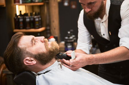 Professional barber using electric razor to shave beard. Regular client at an appointment in barbershop. Confident male customer getting his beard done.の写真素材