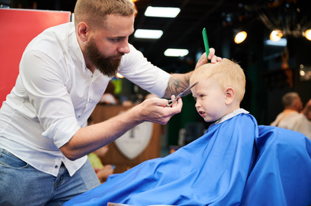 Man barber cutting little boys hair using comb and scissors. Child getting haircut from adult male, likely barber. Professional hairdresser and cute client at modern barbershop.の写真素材