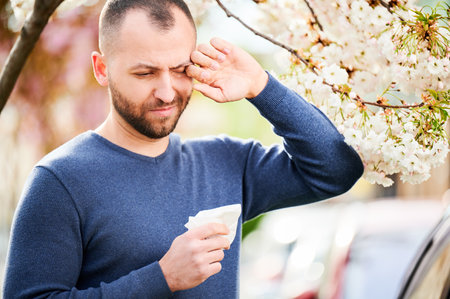 Man allergic suffering from seasonal allergy at spring in blossoming garden at springtime. Man sneezing and feeling itchy eyes in front of blooming tree. Spring allergy concept.の写真素材