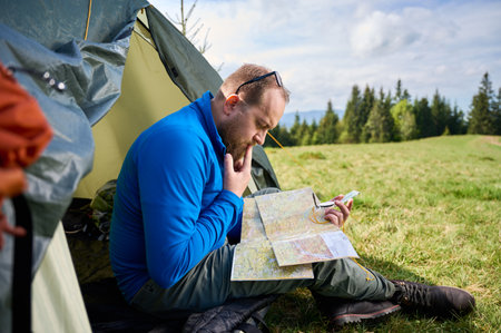 Man sits in tourist tents entrance, deeply focused on map. Tourist holds compass and contemplates his route, surrounded by lush green field with trees and mountains in distance under cloudy sky.の写真素材