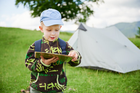 Young boy holds map while hiking in grassy area with tent in background, indicating camping scene. With backpack on shoulders, tourist focused on navigating or planning his route.の写真素材