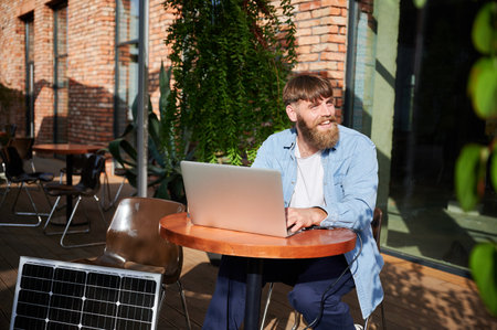 Bearded happy man works on laptop at outdoor round table, with glass of iced coffee. Solar panel nearby, emphasizing sustainable, modern eco-friendly workspace combines technology and sustainability.の写真素材