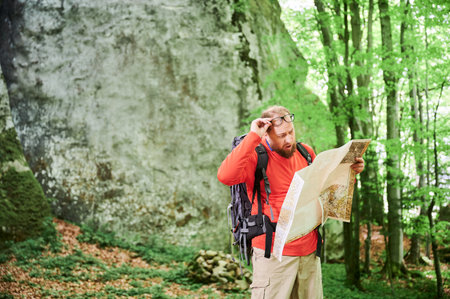 Bearded tourist man looks puzzled with surprised expression, studies map while stands in front of large rock in dense forest. Traveler with backpack and glasses, trying to figure out his direction.の写真素材