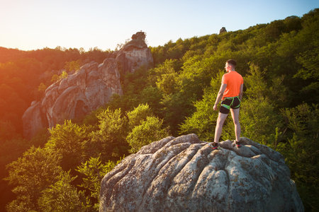 Aerial view of climber standing atop rock formation at Dovbush Rocks in Carpathian mountains, Ukraine. Sun sets, casting warm glow over lush, green forest, distant hills, creating breathtaking view.の写真素材