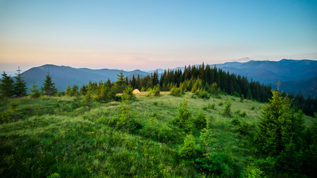Bright yellow tourist tent on grassy hillside, surrounded by lush green pine trees. Breathtaking view of distant blue mountains under clear sky, creating idyllic and serene camping spot.の写真素材