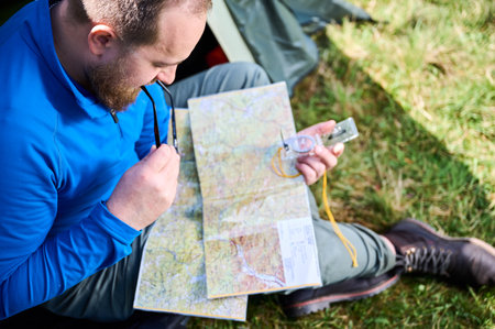 Close up man in glasses closely examines map while holding compass. He sits by his tent, deeply focused on navigation. Lush greenery, suggesting serene outdoor adventure.の写真素材