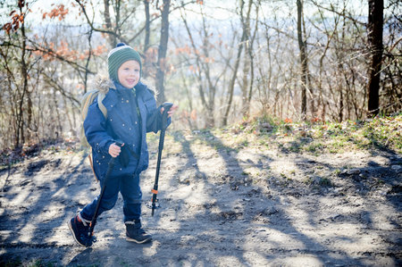 Young child smiling while hiking on forest trail, bundled up in blue winter jacket with fur-lined hood and green striped beanie. Kid holding two trekking poles and enjoying sunny day outdoors.の写真素材