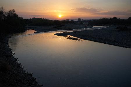 Tranquil sunset graces sky with warm hues of orange and gold, reflecting beautifully off meandering river. Silhouetted trees and distant horizon enhance serene atmosphere of this peaceful landscape.の写真素材
